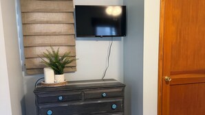 A room with a wooden dresser, a television mounted on the wall, a door, and a patterned rug on the floor. - Rose Haven Coastal Cottage (North Beach)