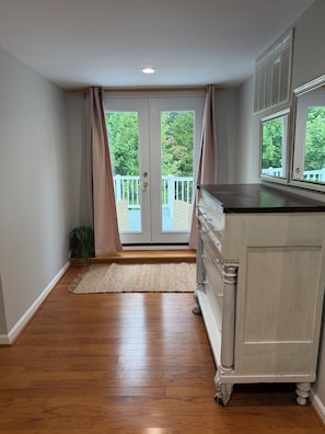 A room with a wooden floor, a white dresser, a window with blinds, and a view of a balcony with greenery.