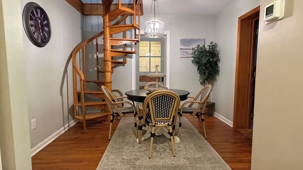 A dining area with a round table, four chairs, a wooden spiral staircase, a clock, a hanging light fixture, and a potted plant.