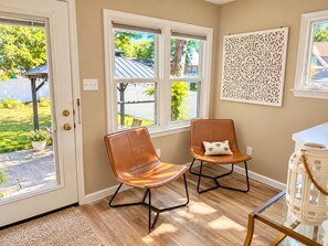 A room with two leather chairs, a white door, and a window with a view of a tree and a building. - Rose Haven Coastal Cottage (North Beach)
