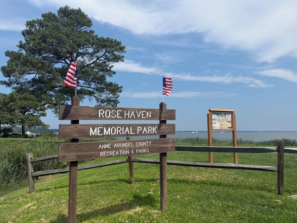 A wooden sign for Rose Haven Memorial Park with two American flags, a grassy area, and a body of water in the background.