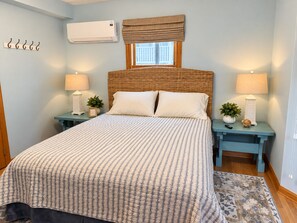A bedroom with a large bed, two bedside tables with lamps, a window with a valance, and an air conditioning unit on the wall.