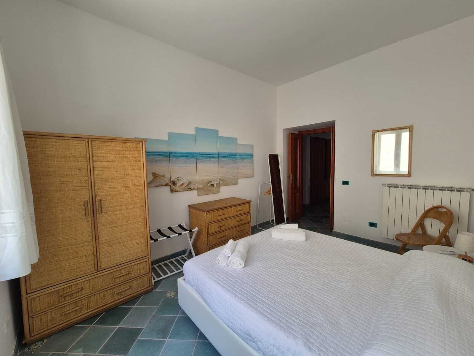 Ein Schlafzimmer mit einem großen Bett, einem Kleiderschrank, einem Schminktisch und einem Bild mit Strandmotiv, sowie einem Sessel.