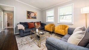 A living room with a dark grey sofa, a yellow armchair, and a grey sofa. There is a coffee table with a vase and a remote control. The room has white walls, a large window with blinds, and a floor lamp. - Stylish 5BR Retreat Near Airport & Downtown (Atlanta)