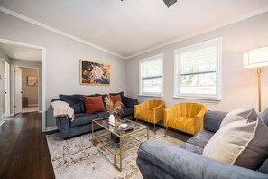 Ein Wohnzimmer mit einem dunkelgrauen Sofa, einem gelben Sessel und einem grauen Sofa. Ein Couchtisch steht dort mit einer Vase und einer Fernbedienung. Der Raum hat weiße Wände, ein großes Fenster mit Jalousien und eine Stehlampe.