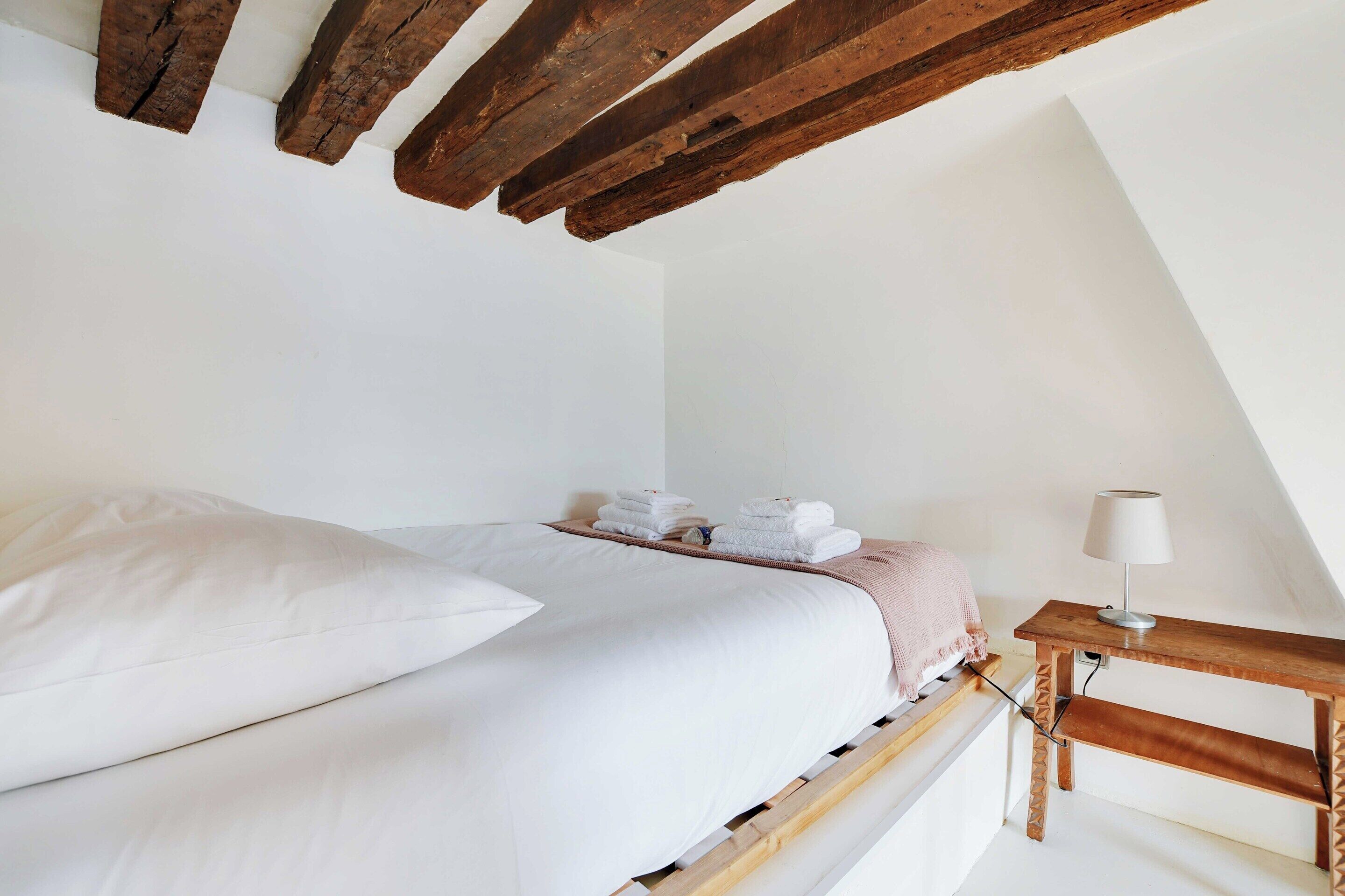 Un dormitorio con cama, almohadas, una mesita de noche con lámpara y una pila de toallas.