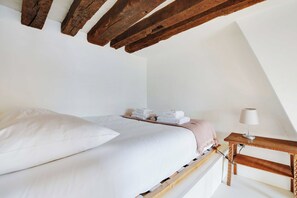 Een slaapkamer met een bed, kussens, een nachtkastje met een lamp en een stapel handdoeken.