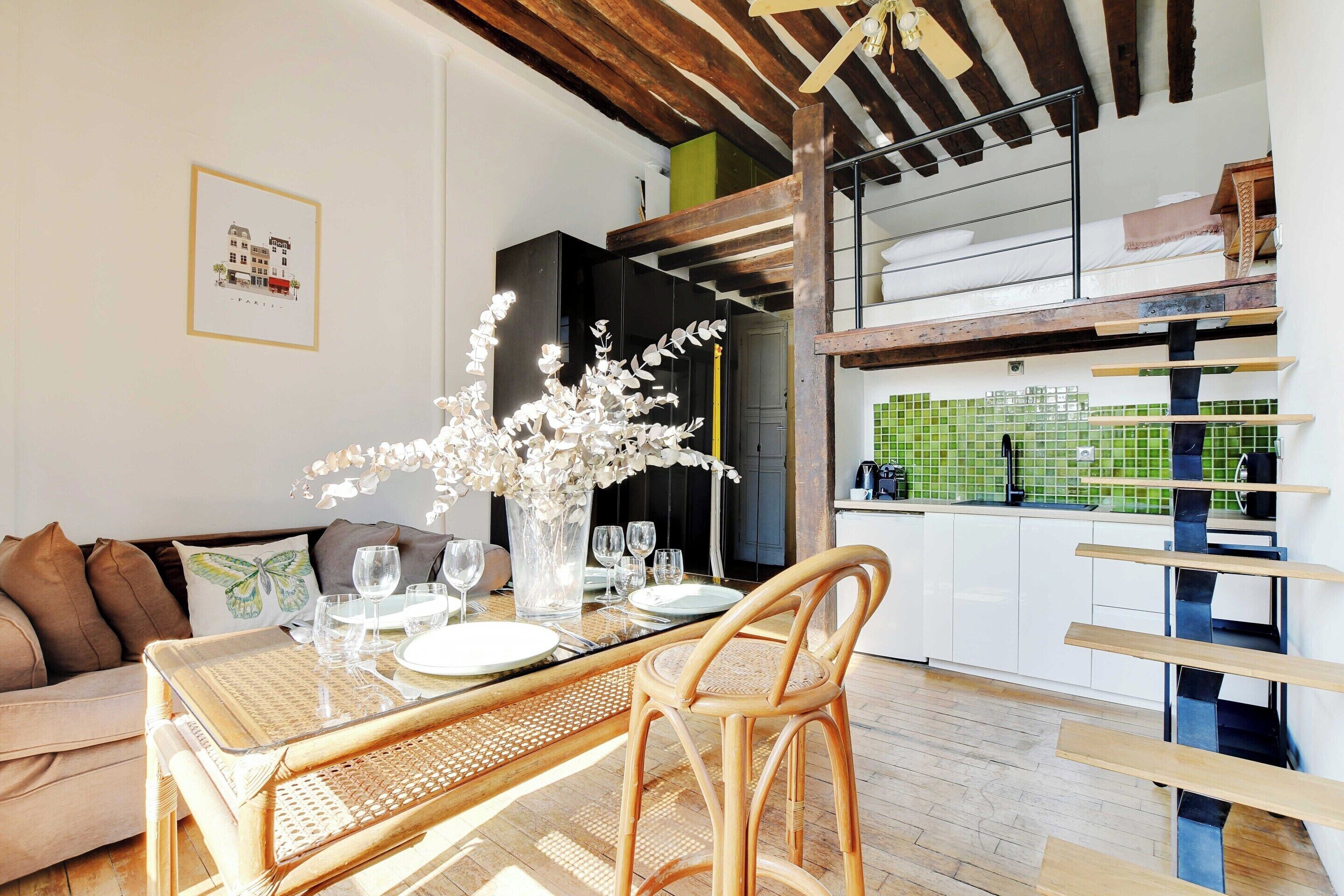 Un comedor con una mesa dispuesta para cuatro personas, una silla de mimbre y un jarrón con flores blancas.
