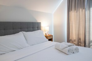 A neatly made bed with white linens and pillows, a grey upholstered headboard, a bedside table with a lamp, and a window with curtains.