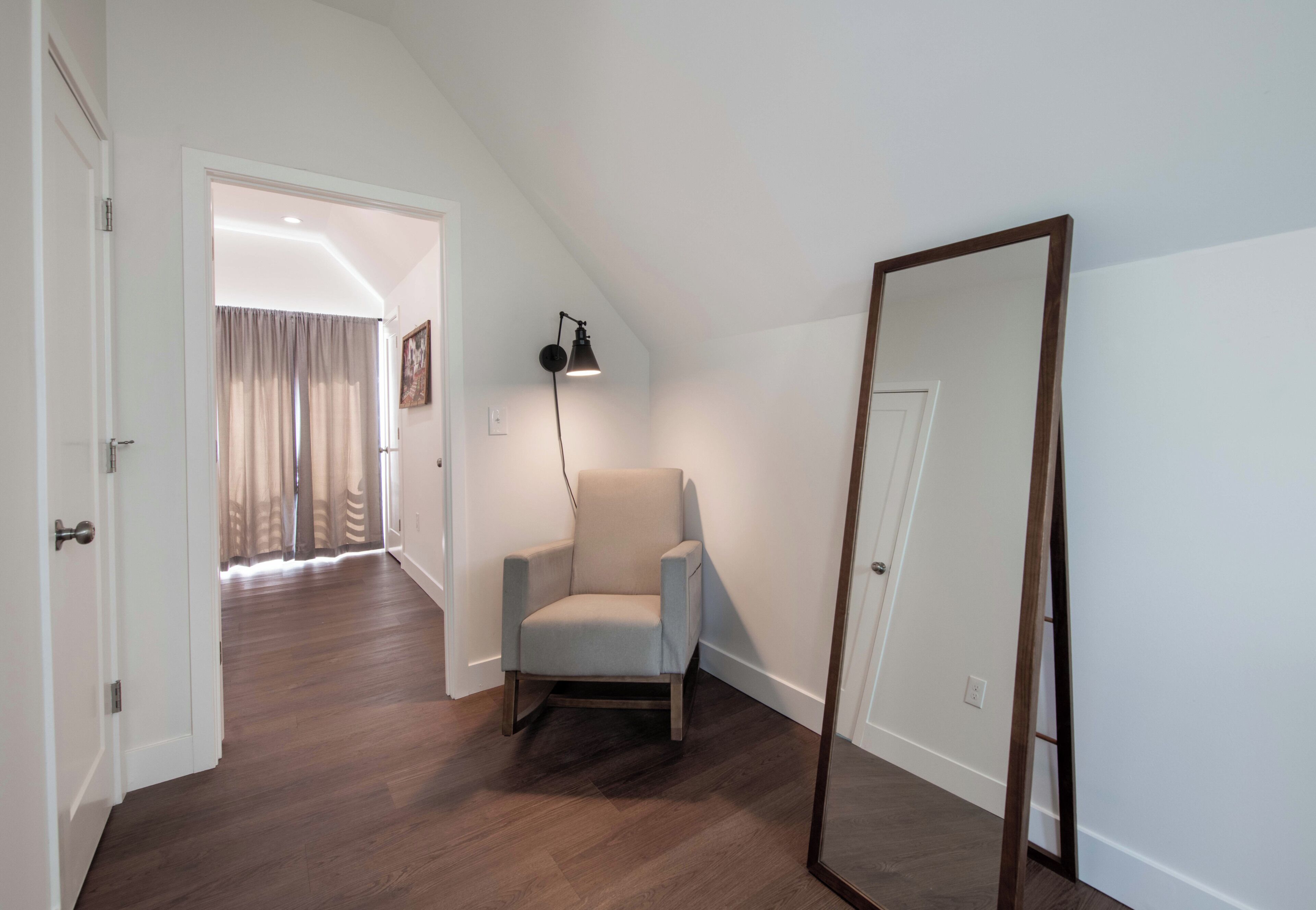 Una habitación con piso de madera, una silla, un espejo y una puerta que da a otra habitación.