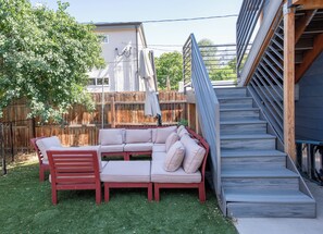 Un patio trasero con una terraza de madera, un banco rojo, un sofá seccional color beige y una escalera que lleva a una zona cubierta.