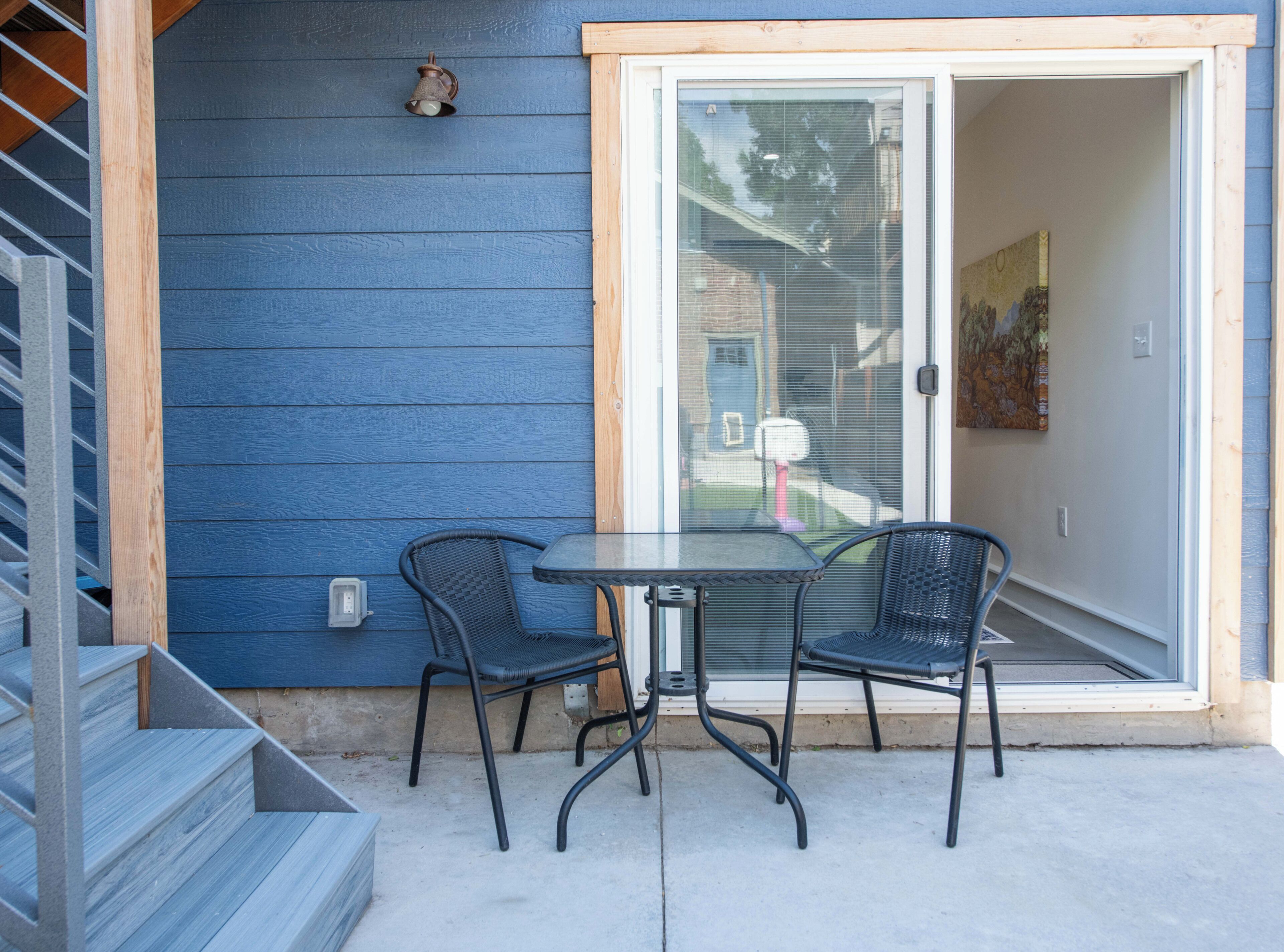 Un patio con puerta de vidrio que da a un espacio cubierto, dos sillas y una mesa pequeña.