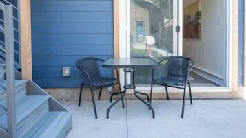 Un patio con puerta de vidrio que da a un espacio cubierto, dos sillas y una mesa pequeña.