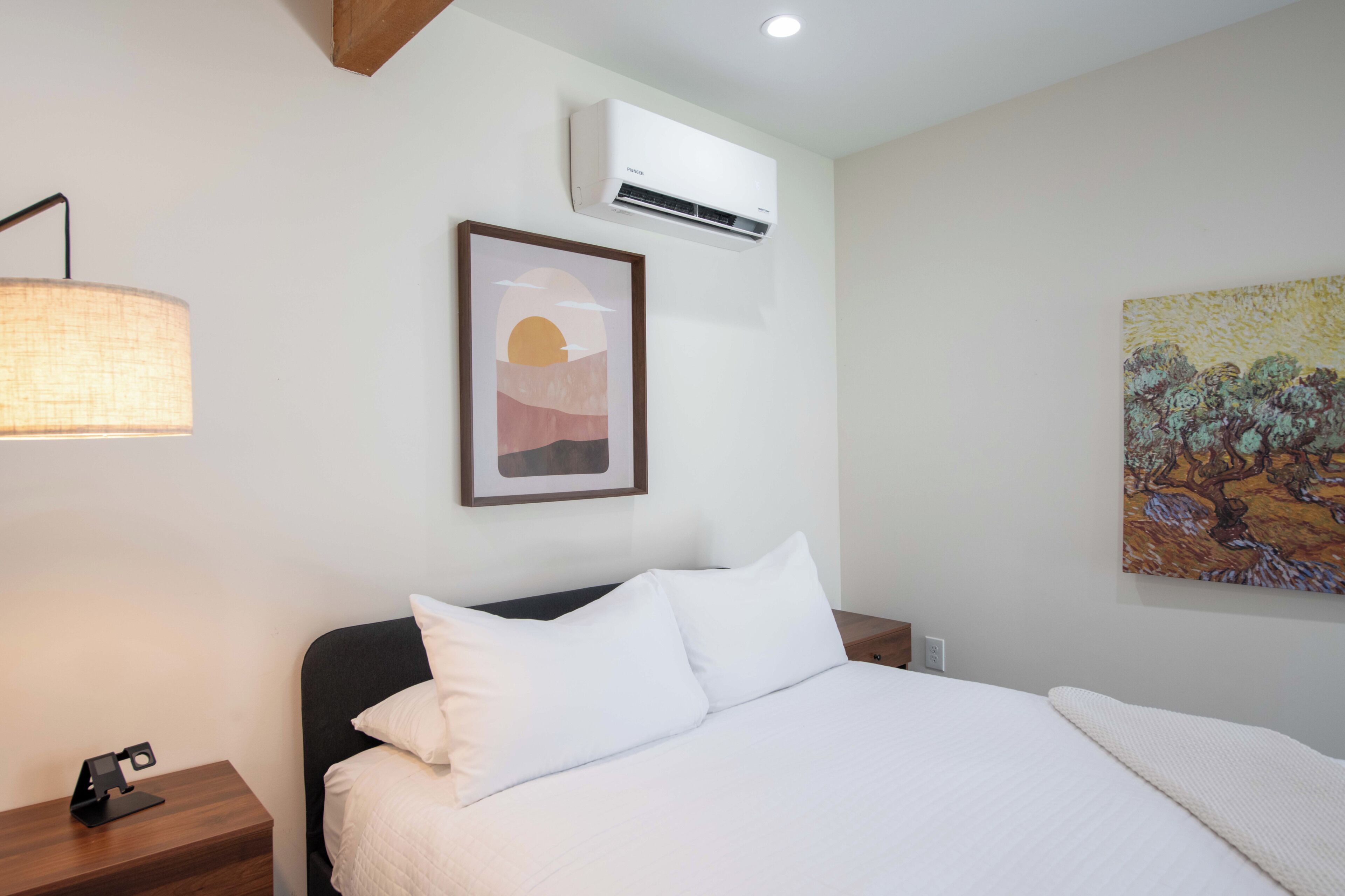Una habitación de hotel con una cama, una mesita de noche, un equipo de aire acondicionado, una lámpara de pared y dos obras de arte en la pared.