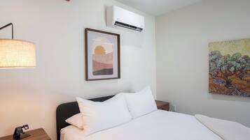 Una habitación de hotel con una cama, una mesita de noche, un equipo de aire acondicionado, una lámpara de pared y dos obras de arte en la pared.