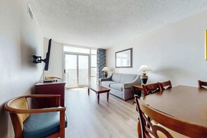 A living room with a sofa, a coffee table, a dining table with chairs, a TV mounted on the wall, and a window with a view. - Carolinian Beach Resort 1030 (Myrtle Beach)