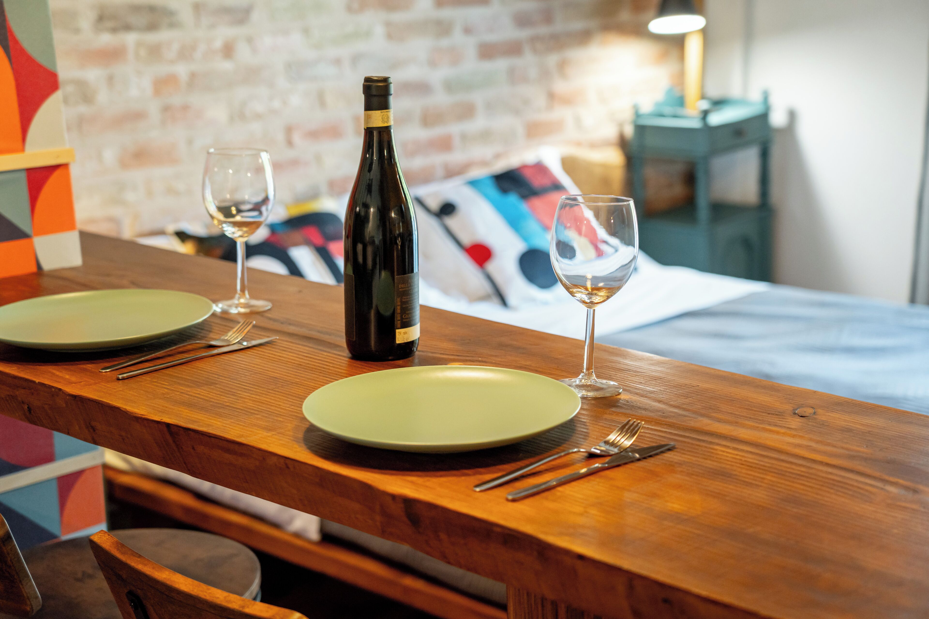 A dining table with a bottle of wine, two glasses, and cutlery, set against a brick wall with a colorful pillow in the background.