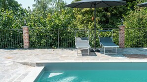 A swimming pool with two lounge chairs and umbrellas, surrounded by a brick wall and a metal fence.