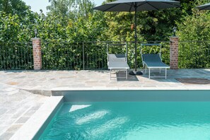 A swimming pool with two lounge chairs and umbrellas, surrounded by a brick wall and a metal fence.