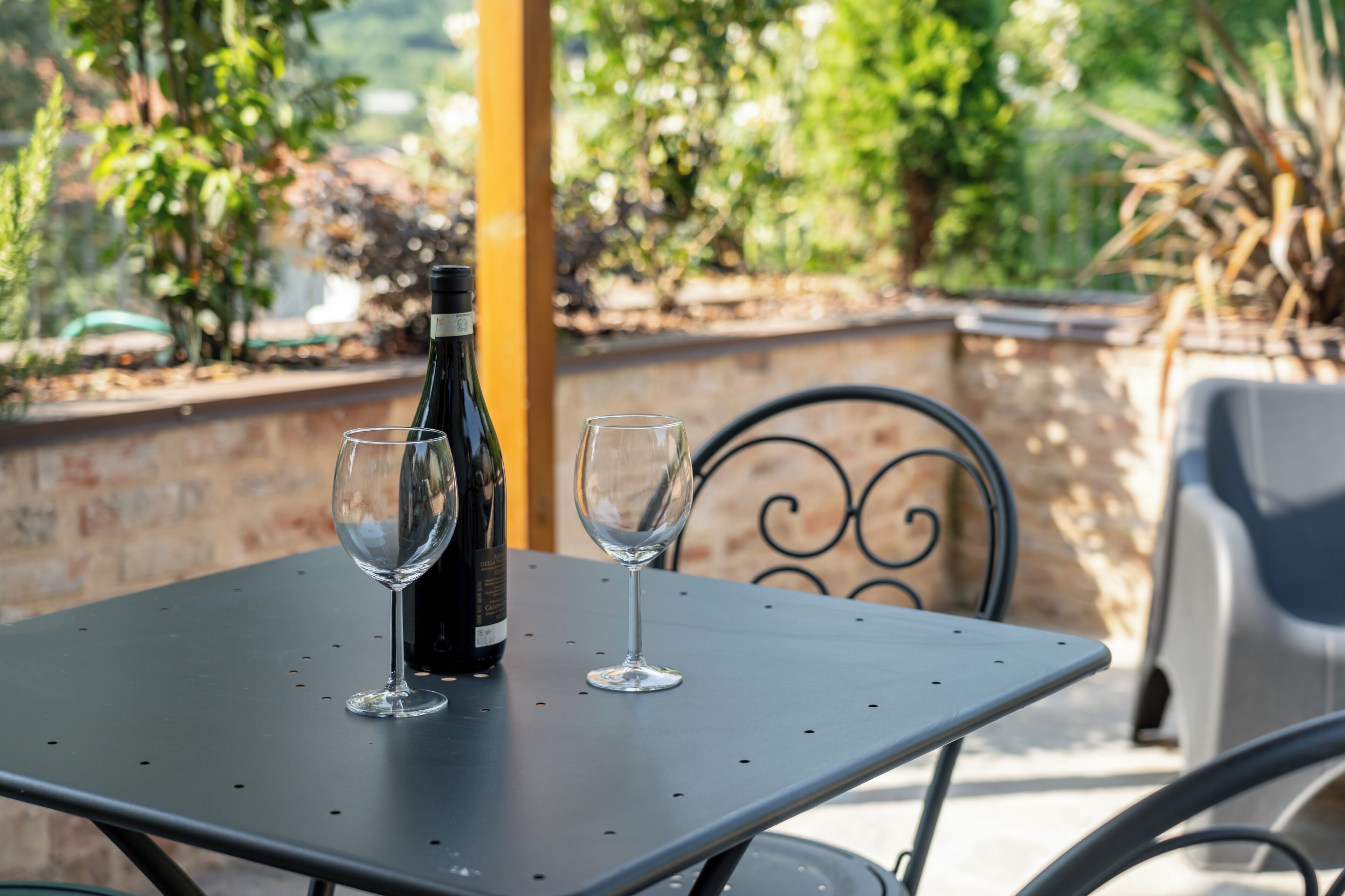 A table with two wine glasses and a bottle, set outdoors with a metal chair and a brick wall in the background.