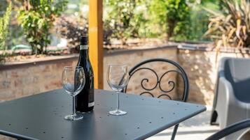 A table with two wine glasses and a bottle, set outdoors with a metal chair and a brick wall in the background.