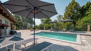 An outdoor swimming pool with lounge chairs and umbrellas, surrounded by a stone patio and greenery.