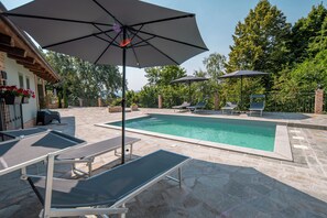 An outdoor swimming pool with lounge chairs and umbrellas, surrounded by a stone patio and greenery.