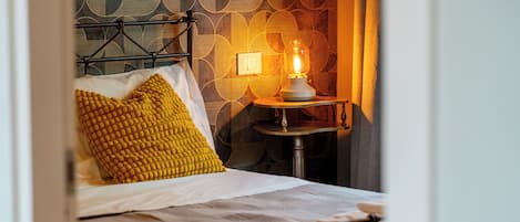 A bed with a metal frame, a white mattress, and two pillows, one yellow and one white. There is a bedside table with a lamp and a wall clock. The wall has a geometric pattern.