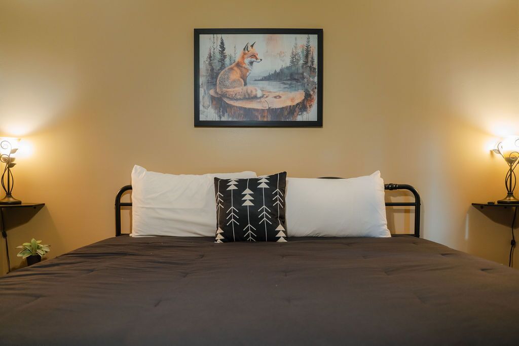 A bed with a dark bedspread, two white pillows, and a black pillow with a tree pattern. A framed painting of a fox by a lake hangs above the bed.