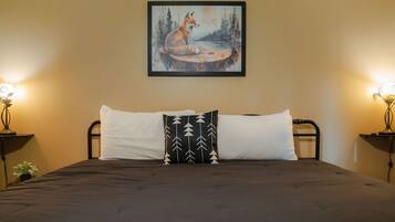 A bed with a dark bedspread, two white pillows, and a black pillow with a tree pattern. A framed painting of a fox by a lake hangs above the bed.