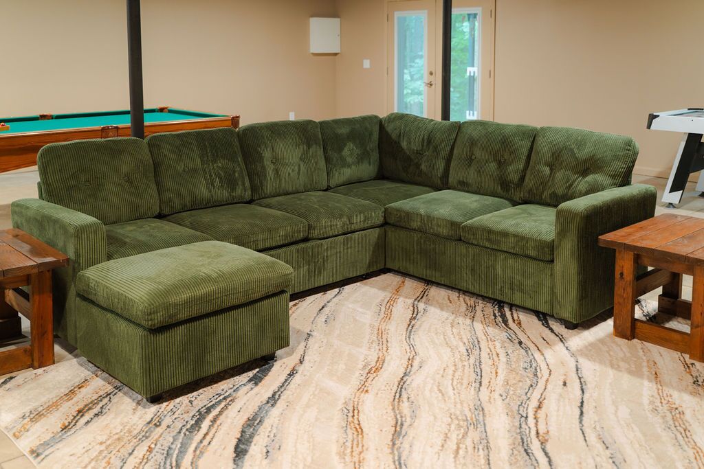 A green sectional sofa with a matching ottoman, a pool table, and a billiards table in a room with a marble-patterned floor.