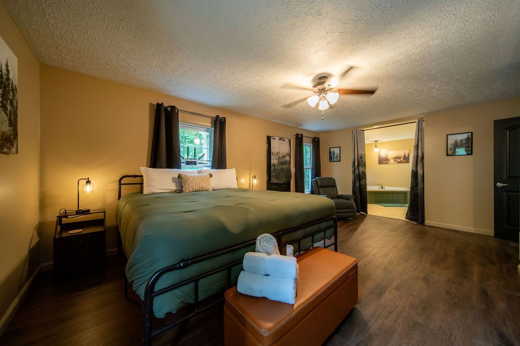A bedroom with a bed, a nightstand, a suitcase, a ceiling fan, and a view of a bathroom.
