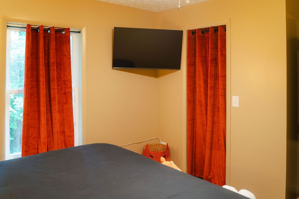 A room with a bed, a flat-screen TV mounted on the wall, and a window with red curtains.