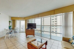 A modern living room with a flat-screen TV, a dining table with chairs, a sofa, and large windows offering a view of the city. - Carolinian Beach Resort 1036 (Myrtle Beach)