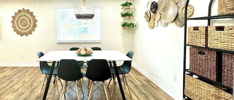 A dining area with a white table, black chairs, a hanging light fixture, and a wall-mounted basket shelf.