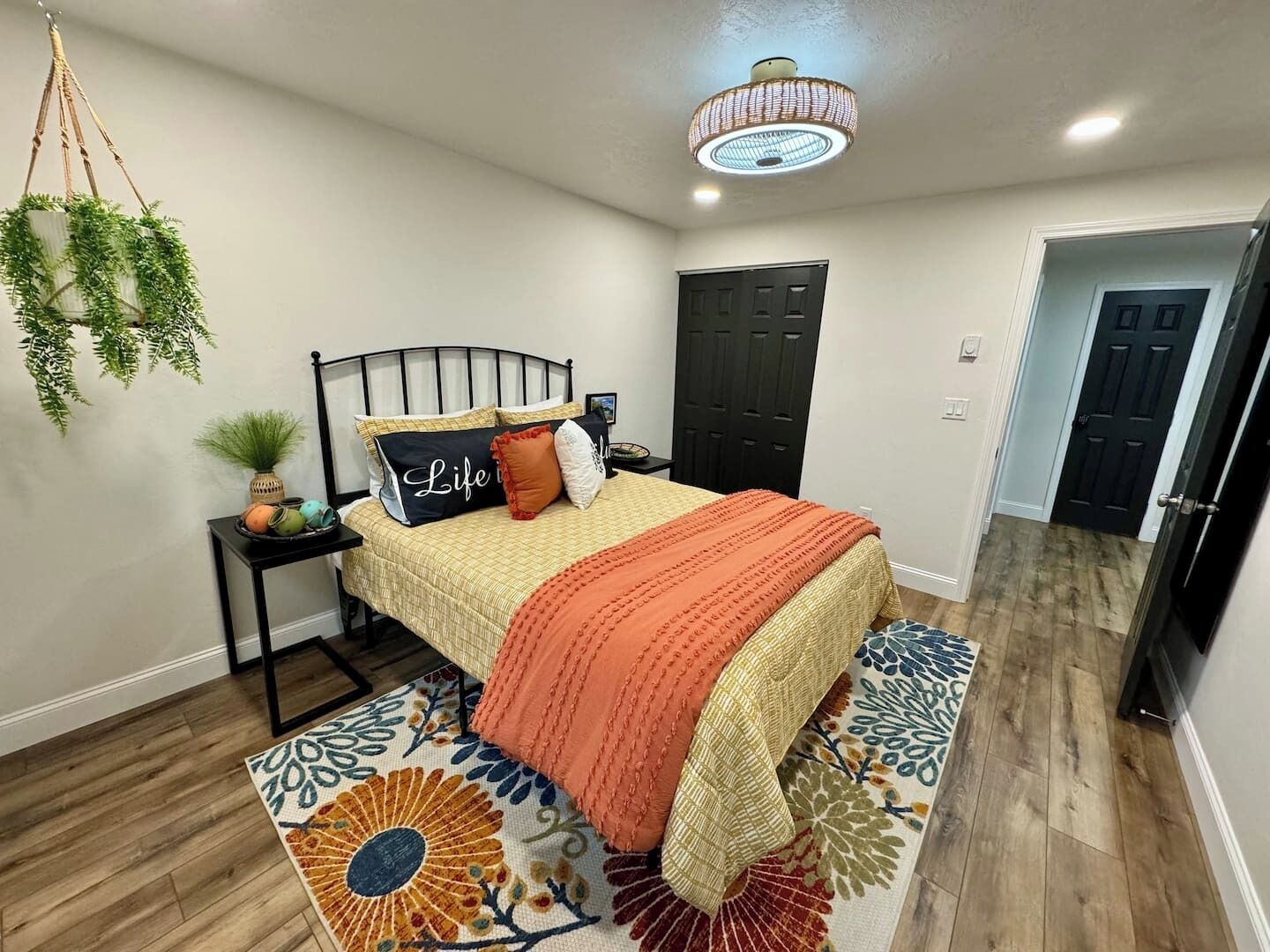 Une chambre à coucher avec un lit, des tables de chevet, une plante suspendue, une porte et un tapis à motifs.