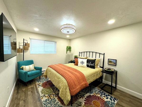 A bedroom with a bed, a chair, a bedside table, a hanging plant, and a ceiling light fixture.