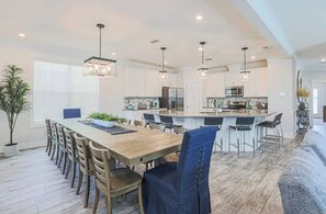 A spacious dining area with a long wooden table, multiple chairs, and pendant lighting. - Lux Family Beach Retreat | Pool Slide • Hot Tub (Hilton Head Island)