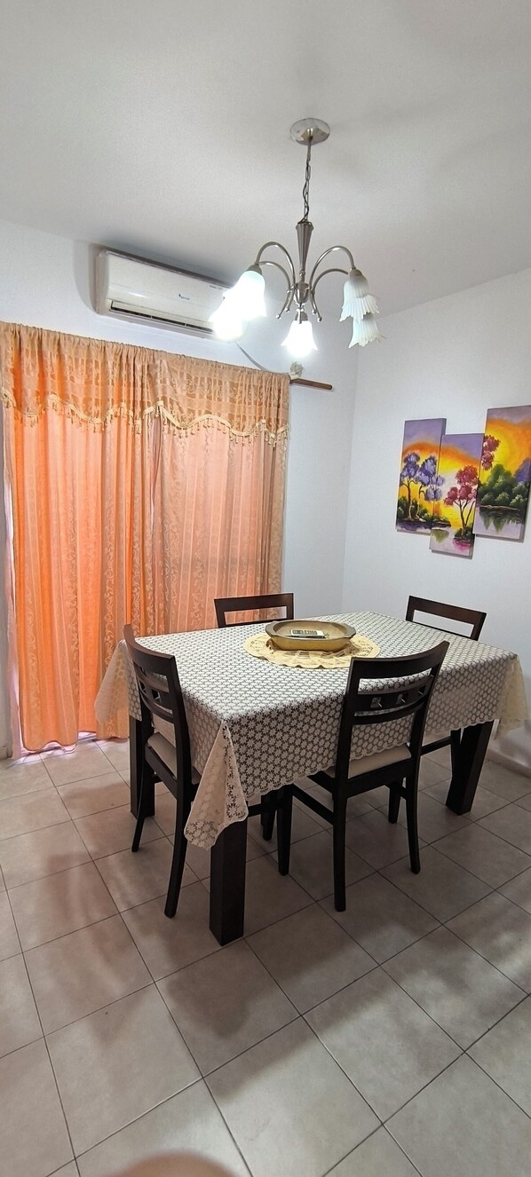 A dining area with a table set for four, chairs, a chandelier, an air conditioning unit, and a window with curtains.