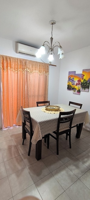 A dining area with a table set for four, chairs, a chandelier, an air conditioning unit, and a window with curtains.