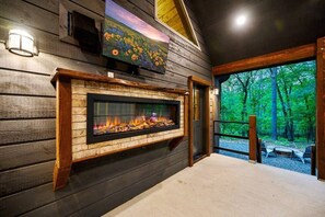 A fireplace with a TV above it, a wooden wall, and a view of a forest through a glass door. - The Guesthouse: Romantic luxury retreat for two with forest views & a soaker tub (Broken Bow)