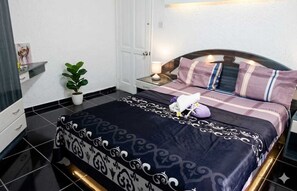 Een net opgemaakt bed met een donker beddekleed, een bedlampje en een kamerplant.