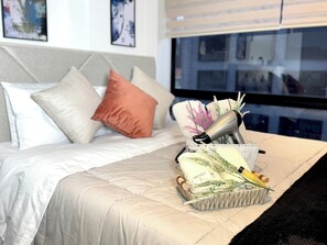 Ein ordentlich bezogenes Bett mit weißer Decke und Kissen, ein Nachttisch mit Fön und Pflegeprodukten sowie ein Fenster mit Blick auf Gebäude.