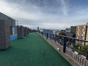 Eine Dachterrasse mit künstlichem Rasen, einem Metallgeländer und Blick auf umliegende Gebäude.