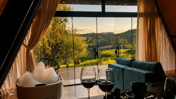 A room with a large window, a sofa, a table with wine glasses and a bottle, and a bowl of ice. - Chalé da Lagoa (Cordilheira Alta)