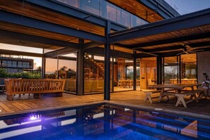 Un espace piscine extérieur moderne, doté d’une terrasse en bois, d’un espace repas clos par des baies vitrées et offrant une vue sur le paysage environnant.