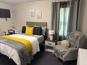 Quarto de hotel com cama, escrivaninha, cadeira, abajur e televisão.