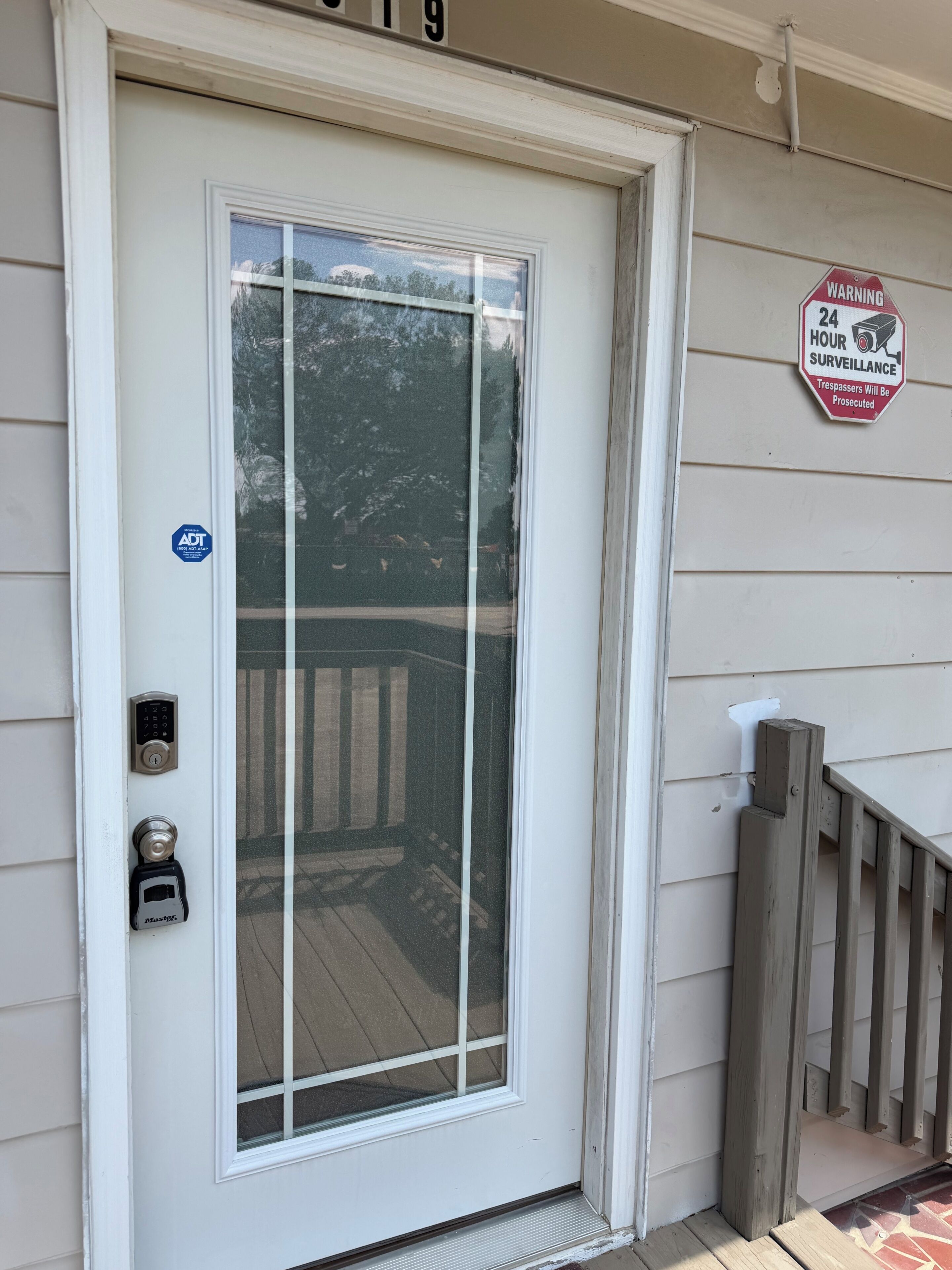 A white front door with a keypad lock, a security camera, and a warning sign for 24-hour surveillance.