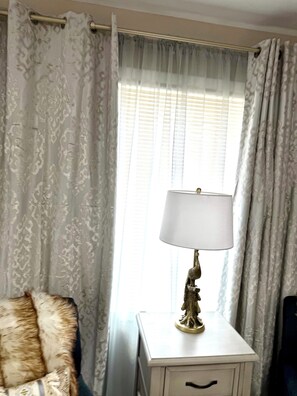 Ein Zimmer mit einem gemusterten Vorhang, einer Lampe auf einem weißen Nachttisch und einem mit Fell bezogenen Sessel.