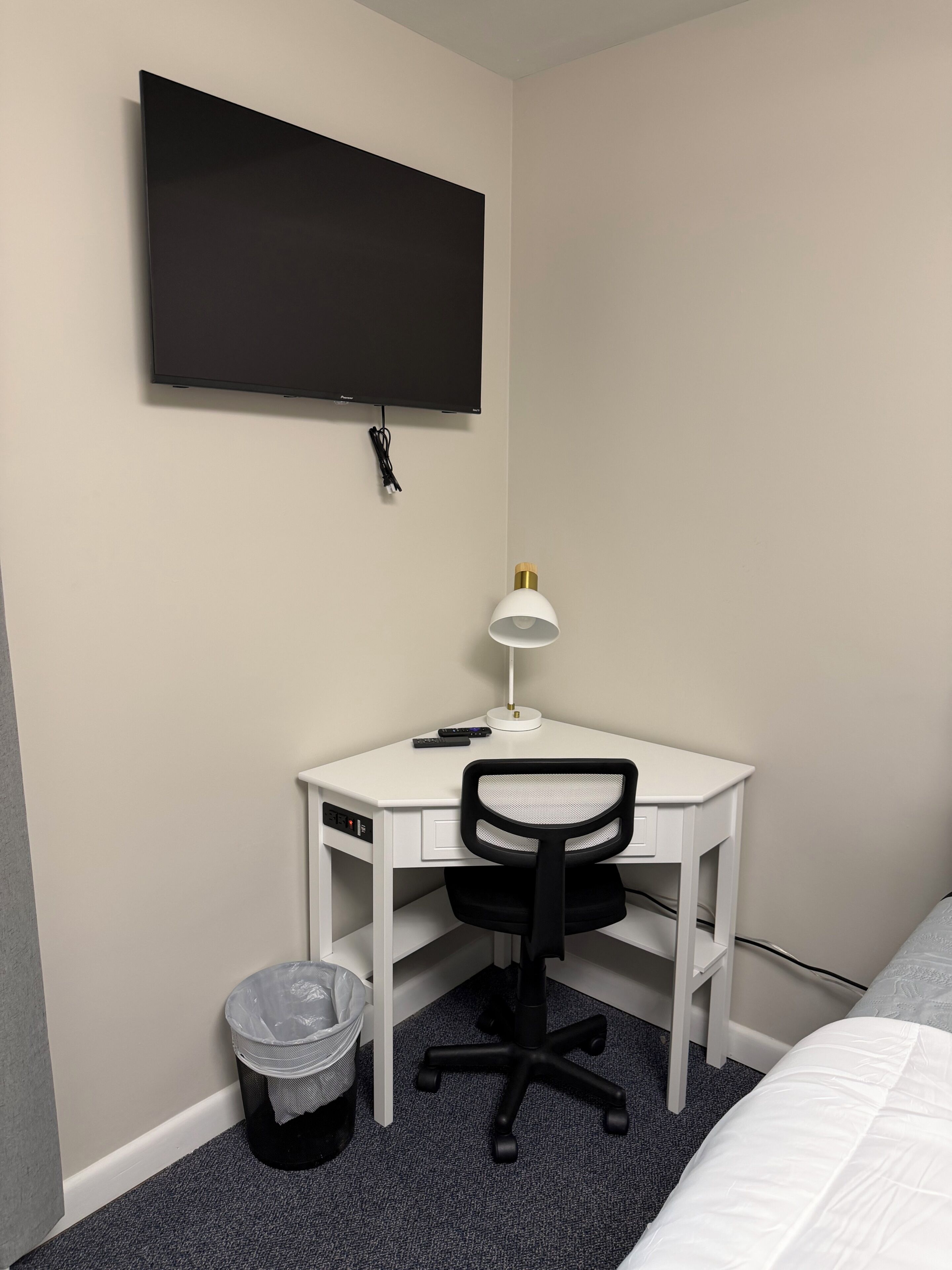 Una habitación pequeña y ordenada, con un escritorio blanco, una silla de oficina negra, un televisor montado en la pared y un cubo de basura.